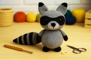 mapache amigurumi tejido a crochet en tonos gris, blanco y negro, sentado sobre una mesa de taller rodeado de herramientas de tejido, ovillos de lana de colores y un entorno artesanal que resalta el detalle y textura del personaje hecho a mano.