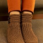 Persona con pantalón naranja claro luce un par de botitas tejidas a crochet en color marrón claro, con detalles en punto relieve y dos botones decorativos en el lateral.