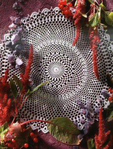 Tapete redondo en crochet de hilo blanco con diseño calado, estilo vintage, sobre fondo color borgoña decorado con flores secas y frutos rojos