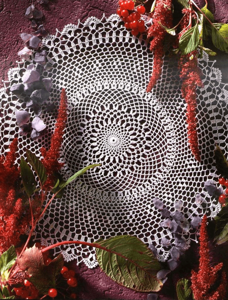 Tapete redondo en crochet de hilo blanco con diseño calado, estilo vintage, sobre fondo color borgoña decorado con flores secas y frutos rojos