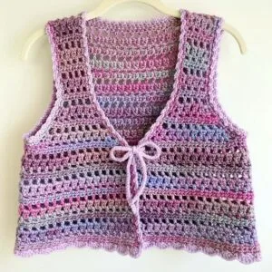 Chaleco tejido a crochet colgado en una percha blanca, confeccionado con hilos en tonos rosa, morado y azul. El diseño presenta un patrón de rejilla a base de puntos altos y cadenas, con un lazo delantero para cierre y un borde ondulado decorativo que resalta la terminación artesanal.