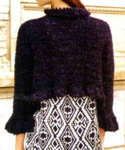 saco corto tejido a crochet con hilo lurex oscuro, de corte corto y mangas largas, lucida de espaldas sobre una falda de estampado geométrico blanco y negro.