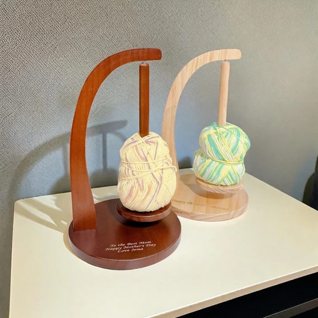 Dos soportes magnéticos de madera para ovillos de hilo, uno en tono oscuro y otro claro, cada uno sosteniendo un ovillo multicolor sobre base personalizada. Ideales para tejer y hacer crochet con comodidad y estilo.