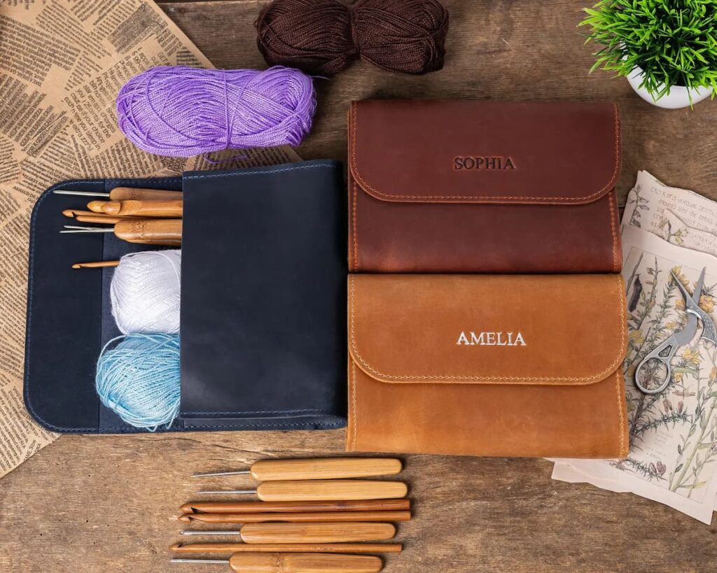 Estuches de cuero personalizados para agujas de crochet, con compartimentos y lanas sobre mesa de madera.