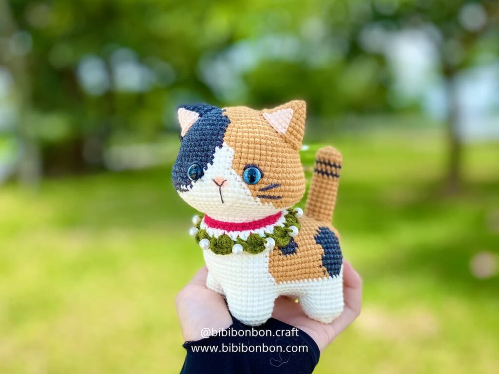 Mano sosteniendo un gato amigurumi tejido a crochet con parches en blanco, negro y naranja, ojos azules y collar rojo, en un entorno verde al aire libre.
