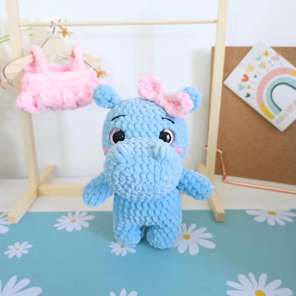 Hipopótama amigurumi tejida a crochet en lana aterciopelada azul, con lazo rosa en la cabeza, junto a un perchero con ropa de crochet.
