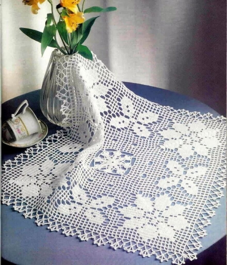 Tapete cuadrado tejido a crochet en color blanco con diseño de flores, colocado sobre una mesa redonda azul junto a un florero de cristal con flores amarillas y una taza decorativa